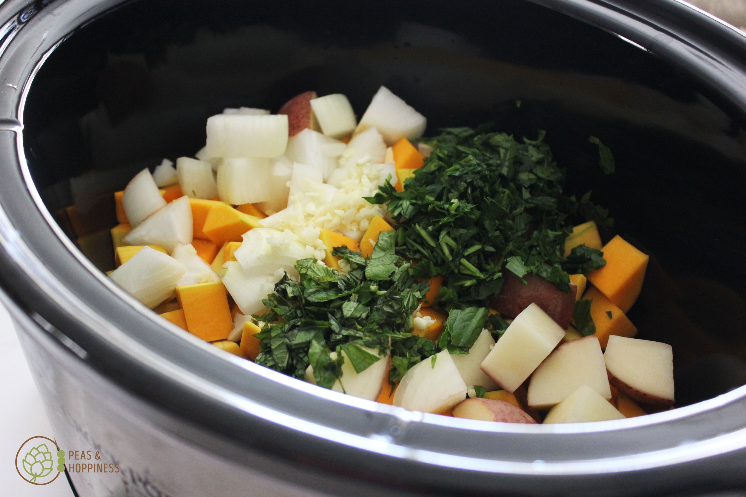 Slow-cooker filled with cubed potato, butternut squash, diced onion, minced garlic, and chopped herbs