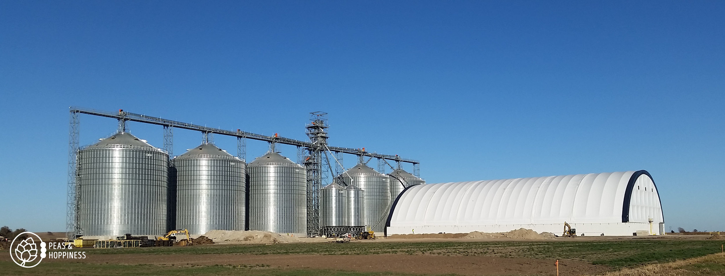 The permanent metal grain storage bins are full, so the the enormous white structure in the foreground was erected for temporary storage this season. See those teeny-tiny back-hoes in front of the building? To give you perspective, those are actuall…