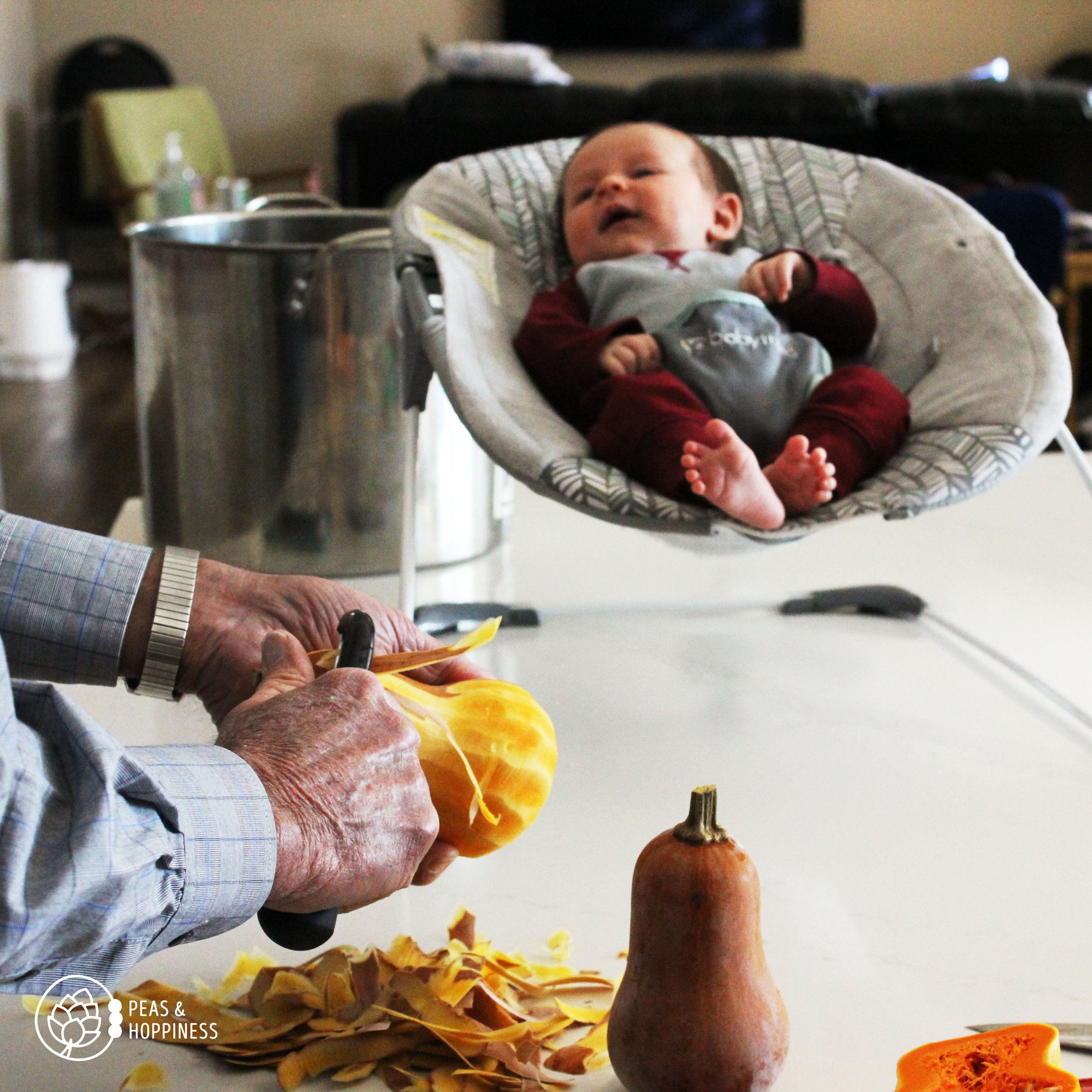 Hands peeling a butternut squash in the foreground with a baby in an infant seat sitting on the counter in the background