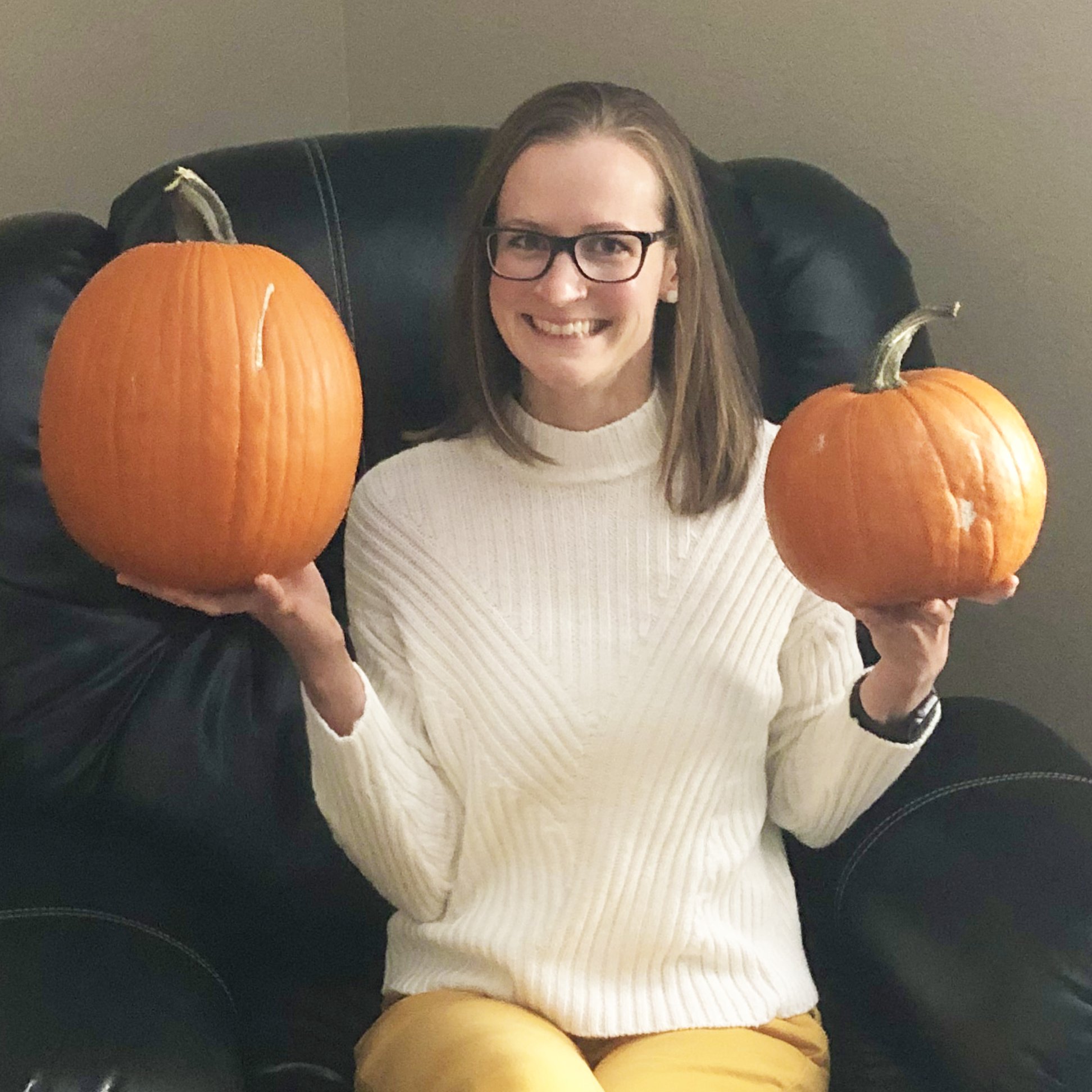 Ann, a woman with shoulder-length brown hair and dark-rimmed glasses sits in a chair and holds two pumpkins of different size