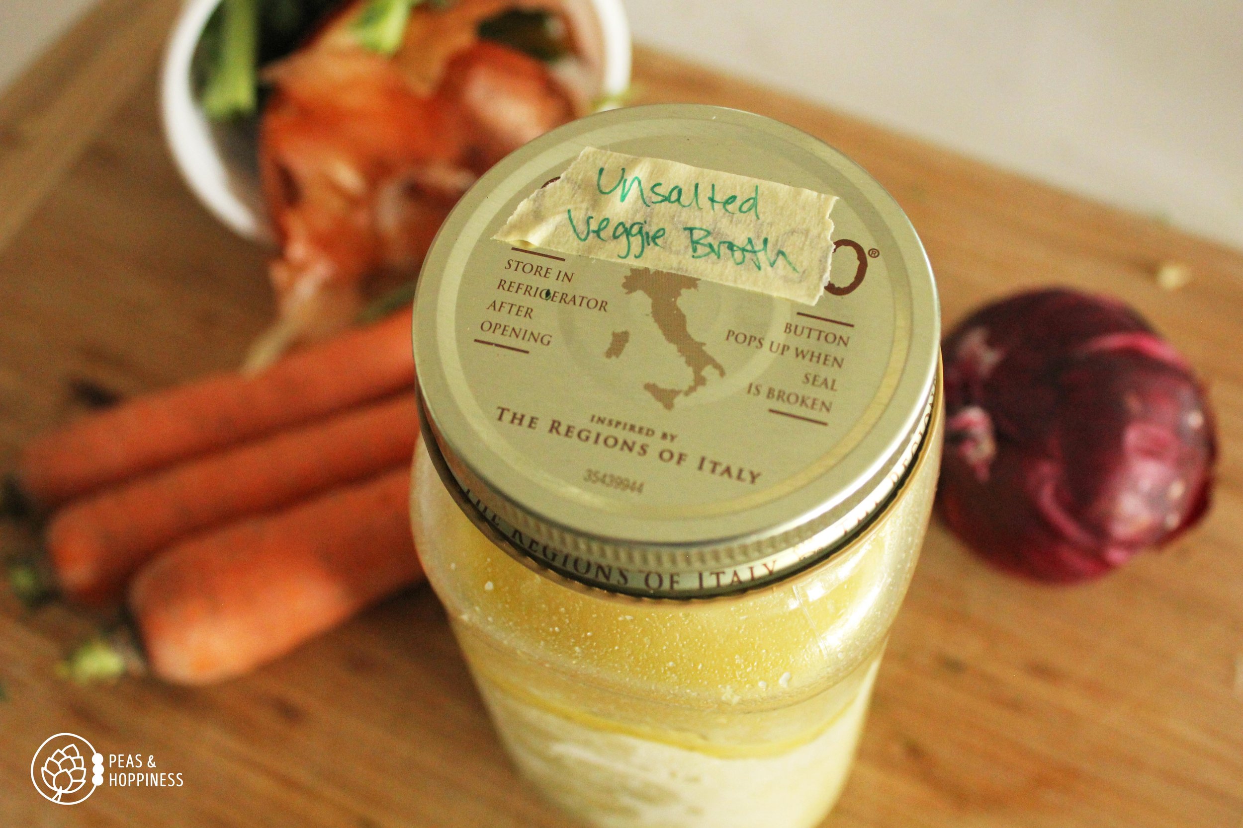 Mason jar with label on the lid that reads "Unsalted Veggie Broth" with vegetables scraps laying on a cutting board in the background
