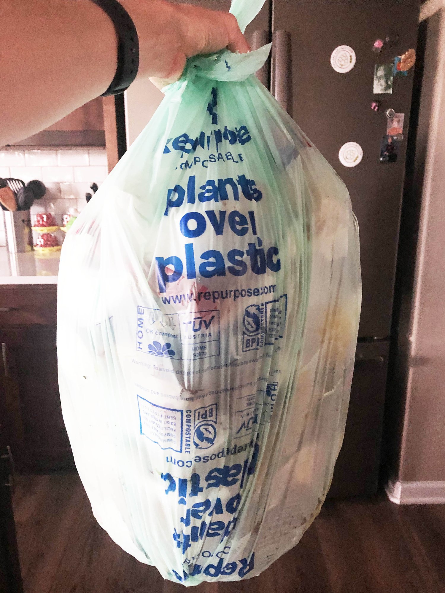Woman's arm holding a trash bag full of trash; trash bag says "plants over plastic" on it