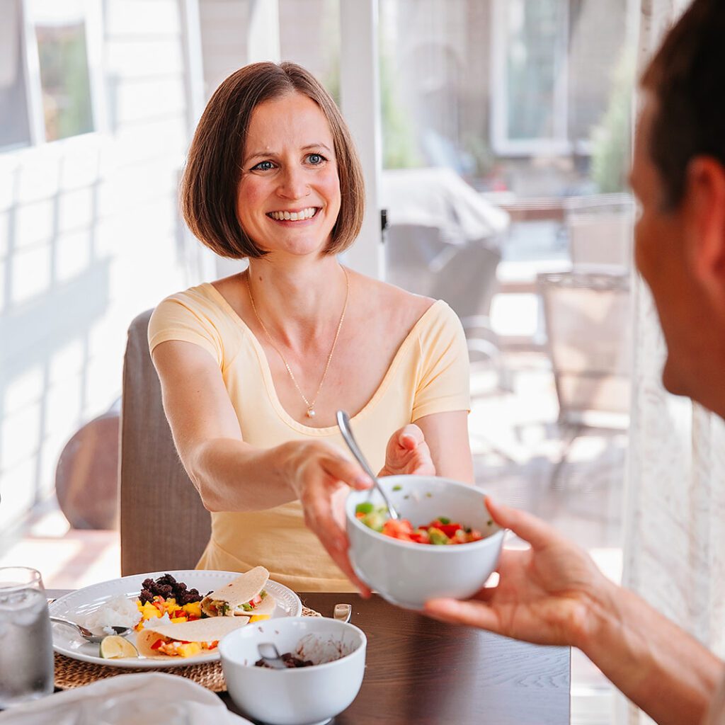 woman with short brown hair handing a bowl of fresh tomato salsa to a man. They are sitting at the dinner table and the woman is smiling.